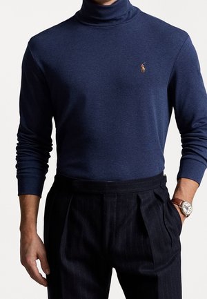 Mann trägt dunkelblauen Rollkragenpullover mit kleinem gesticktem Polo-Spieler-Logo, schwarze Nadelstreifen-Faltenhose und braune Armbanduhr.