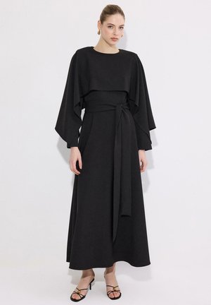 Γυναίκα με μαύρο μακρύ φόρεμα με φαρδιά μανίκια και ζώνη στη μέση, σε συνδυασμό με μαύρα σανδάλια με λεπτές λουρίδες και τακούνι, στέκεται μπροστά σε λευκό φόντο.