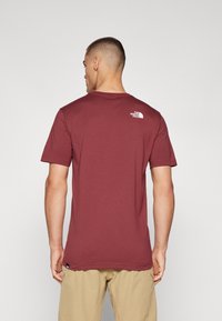 Burgunderfarget kortermet bomulls-T-skjorte med rund hals og hvit logo på øvre bak. Glatt tekstur, avslappet passform, uformell design.
