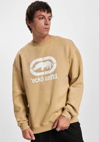 Sudadera beige con un diseño gráfico blanco de un rinoceronte y el texto "*eckō un ltd." en el frente. Material suave, ajuste relajado.