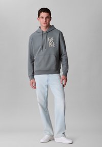 Felpa grigia con cappuccio e tasca frontale, design del logo beige; abbinata a jeans azzurri chiari e sneakers bianche. Texture del tessuto liscia.