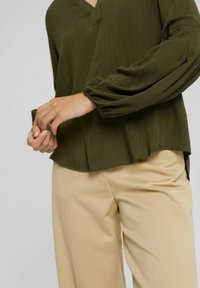 Blouse vert olive avec un col en V, tissu texturé et manches élastiques. Assortie à un pantalon beige, offrant une coupe décontractée et un tombé léger.
