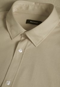 Beige ing texturált anyagból, gombos gallérral és két fehér gombbal az elülső részen. Fekete címke a "Matinique" márkanevével.
