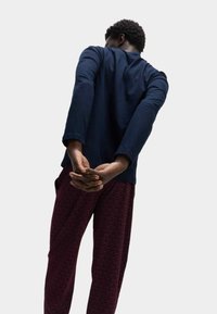 Camisa de manga larga de color marino con dobladillo redondeado, combinada con pantalones color burdeos que presentan un sutil patrón geométrico en toda la superficie. Manos entrelazadas detrás de la espalda.