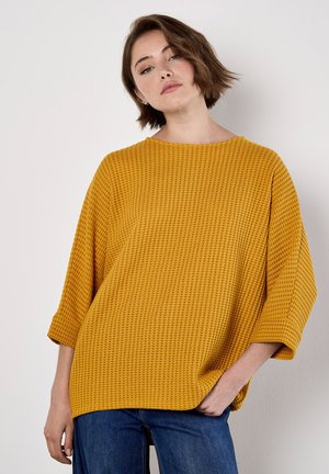 Un haut en tricot texturé jaune avec un décolleté large et des manches trois-quarts, associé à un jean en denim bleu. Un design simple et décontracté.