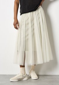 Tüll-Midi-Rock in Off-White, mit geschichteten transparenten Stoffen, elastischem Taillenbund, kombiniert mit beigen Sneakern und gerippten Socken.