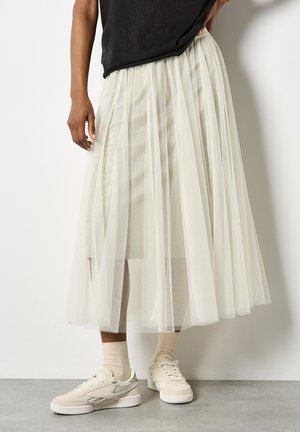 Tüll-Midi-Rock in Off-White, mit geschichteten transparenten Stoffen, elastischem Taillenbund, kombiniert mit beigen Sneakern und gerippten Socken.