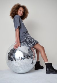 Palla da discoteca con superficie a mosaico riflettente; persona con maglietta grigia, gonna di paillettes e stivaletti neri alla caviglia, seduta sulla palla.