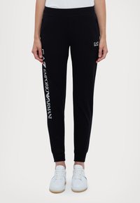 Svarta sweatpants med ribbade muddar, med vit "EA7 Emporio Armani" logotyp på vänster ben; tillverkade av mjukt material. Bärs med vita sneakers.