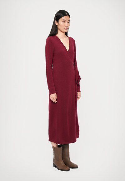 Burgundy wrap dress with long sleeves, deep V-neck, and bow accent at waist. Paired with brown heeled ankle boots. Smooth knit texture.