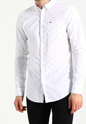 Homme portant une chemise blanche à petits motifs noirs et un pantalon noir, debout devant un fond uni.