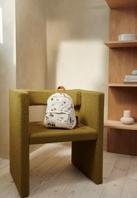 Obdélníková zelená židle s texturovaným potahem vedle světlé dřevěné police. Na židli leží bílý batoh s potiskem zvířat.