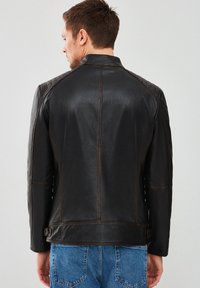 Chaqueta de cuero negra con un respaldo texturizado, acentos acolchados en los hombros y costuras en contraste. Lleva puesta sobre unos jeans de denim, mostrando un diseño ajustado.