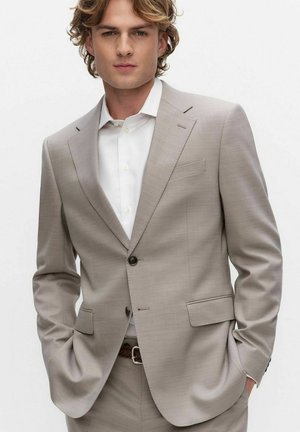 Jonge man draagt een lichtbeige pak en wit overhemd, met handen in de zakken, staand tegen een effen witte achtergrond.