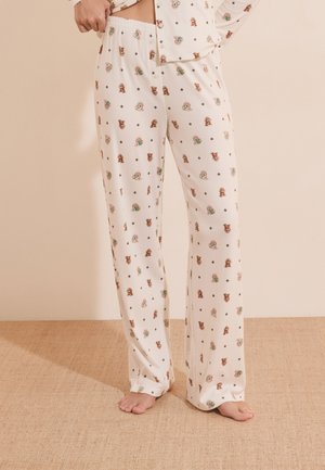 Persoon die crèmekleurige pyjamabroek draagt met kleine teddybeer- en bloemenpatronen, staat blootsvoets op een gestructureerde vloer.