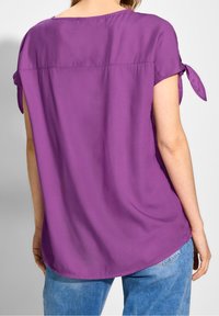 Chemise violette à manches courtes avec une coupe décontractée, dotée de liens en satin aux manches et d'un tissu lisse et léger. Le dos est légèrement arrondi.