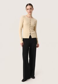 Ribbade beige koftan med rund halsringning, prydd med åtta frontknappar och en kort design, ihop med svarta vida byxor.