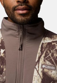 Fleecejacke in Braun und Creme, mit hochgeschlossenem Kragen, durchgehendem Reißverschluss und Columbia-Logo an der unteren rechten Seite. Texturiertes Material und Designdetails sichtbar.