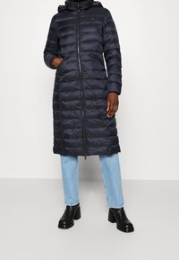 Lång marinblå pufferjacka med huva, med en dragkedja fram, sydda sektioner och sidofickor. I kombination med ljusblå jeans och svarta klackstövlar.