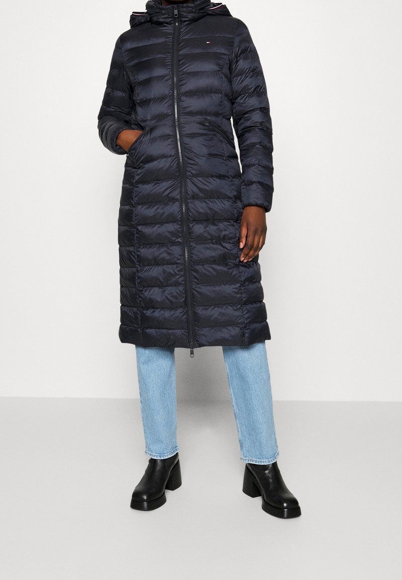 Lång marinblå pufferjacka med huva, med en dragkedja fram, sydda sektioner och sidofickor. I kombination med ljusblå jeans och svarta klackstövlar.