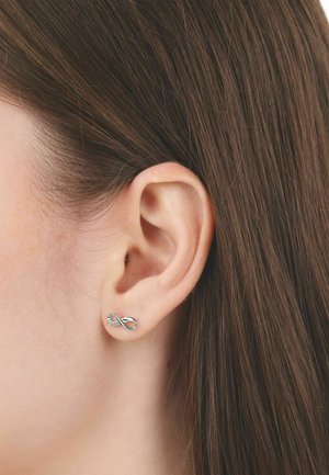 Nahaufnahme vom Ohr einer Frau mit glattem braunem Haar, die einen kleinen silbernen Ohrstecker in Form eines Unendlichkeitssymbols trägt.