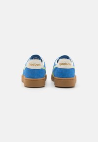 Sneaker in camoscio blu con accenti verdi e suola in gomma marrone. Presenta un tallone bianco con il logo "Reebok" in oro.
