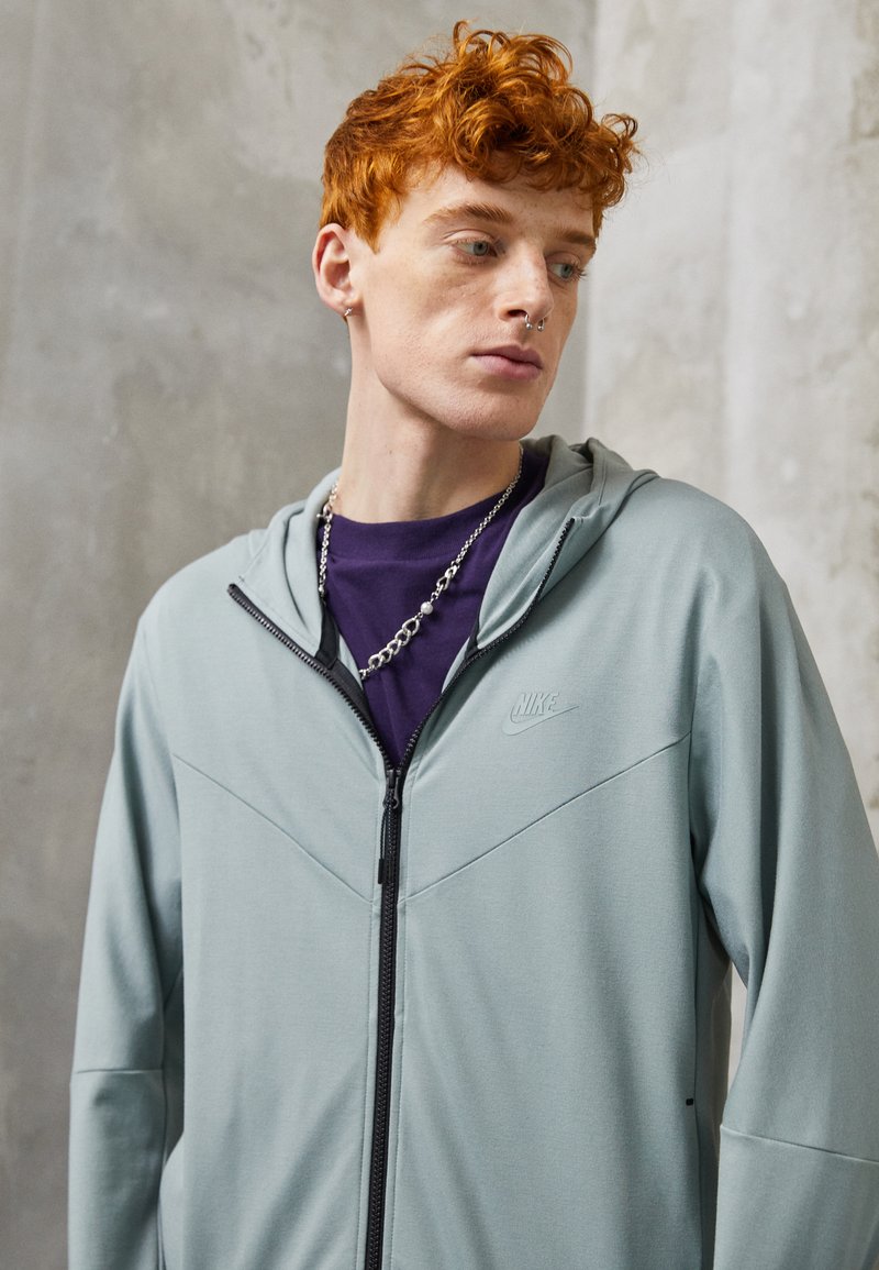 Haine à capuche zippée vert clair avec une capuche, présentant un design texturé et un logo Nike subtil. Portée sur une chemise violette, avec un détail en chaîne argentée.