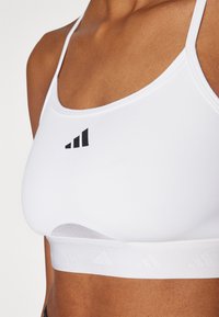 Sujetador deportivo blanco con tirantes ajustables, detalle del logo negro en la parte frontal y una banda blanca texturada en la parte inferior. Diseño casual y funcional.