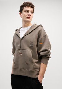 Chaqueta con cremallera color caqui que presenta animales bordados, un bolsillo delantero y puños acanalados. Confeccionada en una tela suave y con un corte holgado.