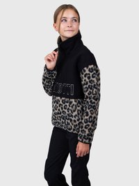 Młoda osoba zapinająca zamek błyskawiczny czarnej polarowej kurtki z rękawami w leoparda i napisem "QUOTTI", ubrana w czarne spodnie.