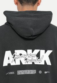 Sudadera con capucha negra que presenta un gran gráfico blanco con el texto "ARKK" y "Innovación Incluida" en la parte posterior. Textura de tela suave.