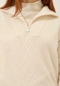 Kremowy pulower z pół-zamkiem przy szyi, z prążkowaną teksturą i diamentowym wzorem w okolicy klatki piersiowej. Miękki materiał dzianinowy.