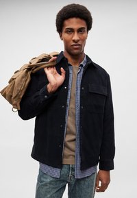 Crna jakna od antilopa sa kopčama na drukere, nošena preko plave karirane košulje i bež majice, u kombinaciji s plavim trapericama i smeđom torbom u ruci.