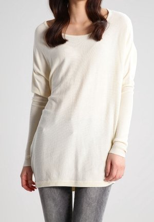 Maglione beige morbido con scollatura ampia, maniche lunghe e texture a costine. Abbinato a jeans grigi, presenta un orlo leggermente allungato.