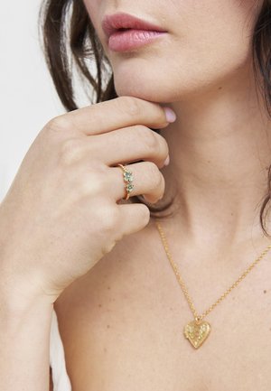 Naisteras puudutab lõuga, kandes kuldset sõrmust siniste lilledega ja kuldset südamekujulist ripatskaelakeed paljal nahal.