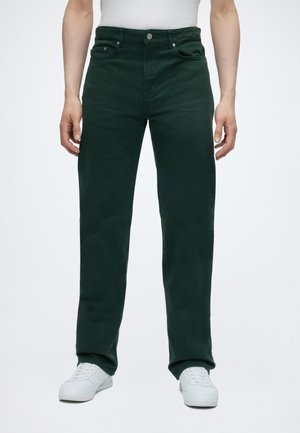 Uomo che indossa pantaloni dritti verde scuro e sneakers bianche, in piedi davanti a uno sfondo bianco semplice.