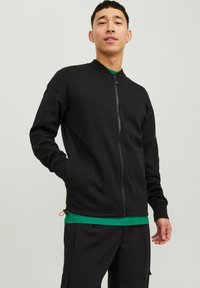 Črna zip-jopica s stoječim ovratnikom, izdelana iz gladkega blaga, z dvema sprednjima žepoma in diskretno oranžnimi detajli ob robu.