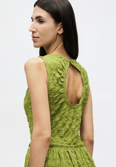 Γυναίκα με ίσια σκούρα μαλλιά, που φοράει ένα πράσινο φόρεμα χωρίς μανίκια με υφή, ανοιχτή πλάτη και κλείσιμο με κουμπί στον λαιμό.