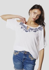 Blusa blanca de manga corta con hombros descubiertos, decorada con bordado floral azul marino a lo largo del escote y tela con textura sutil.