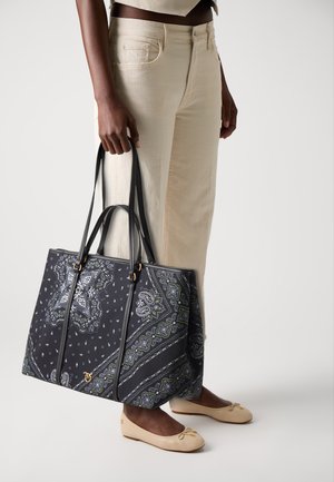 Persoon in crèmekleurige broek en beige flats die een grote zwarte tote bag vasthoudt met een wit en groen paisleypatroon.
