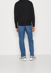 Schwarzes Langarmshirt, blaue Jeans und weiße Sneakers mit schwarzen Akzenten. Das Shirt hat einen gerippten Halsausschnitt und Bündchen.
