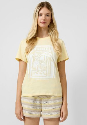 Junge Frau mit langen blonden Haaren, die ein blassgelbes Tahiti-Grafik-T-Shirt und gestreifte Strickshorts trägt, steht vor einem schlichten Hintergrund.