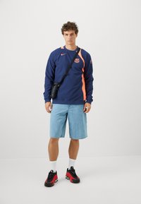 Marineblå pullover med orange detaljer, logo på brystet, parret med lyse denimshorts og sorte/røde sportssko. Inkluderer en lille sort taske.