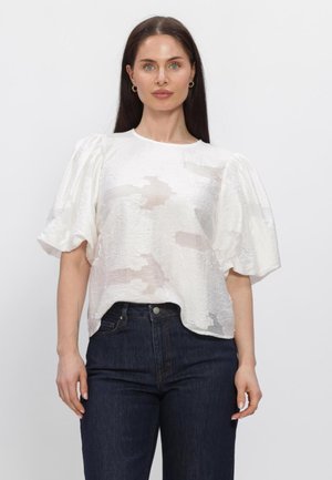 Vrouw met lang donker haar, gekleed in een witte blouse met pofmouwen en een donkere broek met hoge taille, staande tegen een effen lichte achtergrond.