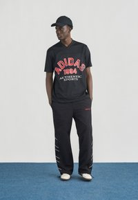 Νέος άντρας με μαύρο καπέλο Adidas, μαύρη αθλητική φανέλα Adidas με κόκκινο κείμενο, μαύρο παντελόνι Adidas με λευκές λεπτομέρειες και λευκά αθλητικά παπούτσια, στέκεται σε μπλε χαλί.