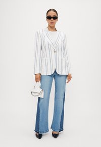 White striped blazer with a single button, paired with flared blue jeans. The outfit features a white handbag and black sunglasses.