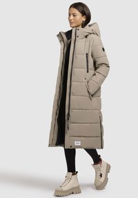 Beige lang pufferjakke med hætte, lynlås foran, sidelommer og kontrasterende sorte detaljer. Tykke quiltede mønstre og glat stof.