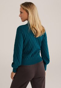 Knitted teal sweater with textured chevron pattern, ribbed cuffs and hem, paired with dark brown pants. Model shown from the back.