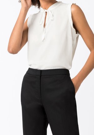 Femme portant un chemisier blanc sans manches avec un détail de nœud au col et un pantalon noir sur mesure, une main dans la poche, sur fond blanc.