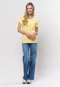 Kollane puuvillane T-särk ümmarguse kaelusega, kombineeritud helesiniste lainerõngastega. Modell seisab vabas poosis, käed tal üleval talje juures.